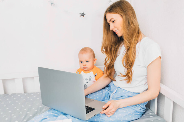 Egy fiatal anya pizsamában, egy kisgyerekkel, az ágyon ülve, hordozható laptoppal. A karanténban végzett távmunka fogalma - Fotó, kép