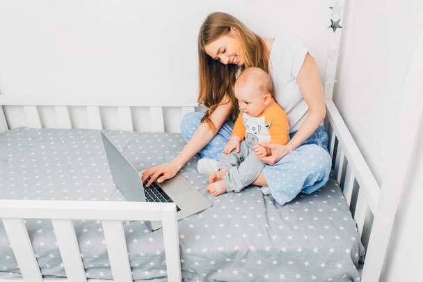 Mladá matka v pyžamu s malým dítětem, sedící na posteli s přenosným laptopem. Koncept vzdálené práce v karanténě - Fotografie, Obrázek