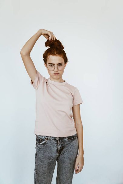 a young schoolgirl in glasses for vision twisted her hair into a bun on her head and holds it with her hand, while blowing her lips and expressing insult - 写真・画像