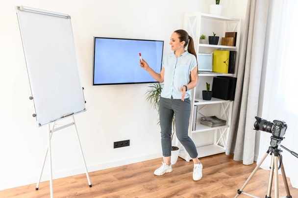A young woman is online teacher, tutor, mentor. A woman in smart casual is recording video classes, she is using flipchart and screen for explanation - Zdjęcie, obraz