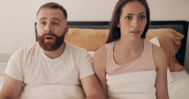 Cute young couple oversleep and looking at each other with big eyes and than falling back on bed . Man and woman covering blanket lying on the pillows and waking up with screaming. - Materiał filmowy, wideo