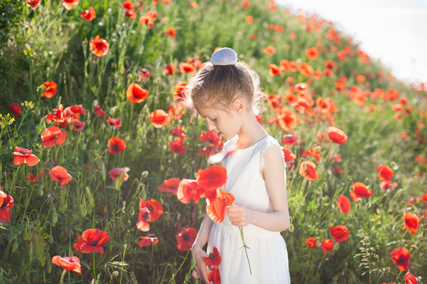 beautiful little caucasian girl in white dress holding poppy red in green summer field - Foto, Imagem