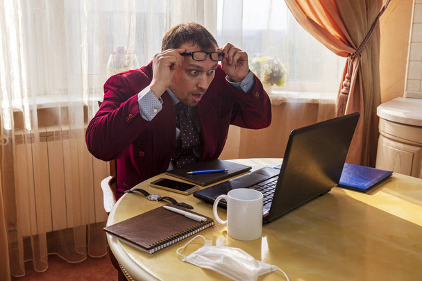 Remote work, Sitting at the kitchen table, a young man in a business suit and shorts holds a meeting, discussing problems on social networks with business colleagues, terrified of information, glasses on the forehead - Photo, Image