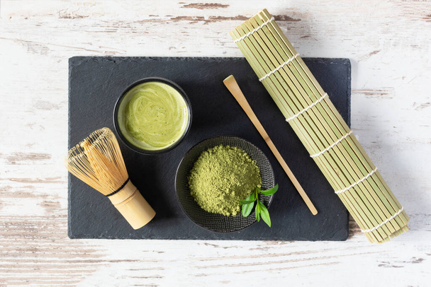 Green matcha tea drink and tea accessories on black board on white wooden table top view. Japanese tea ceremony concept. Chashaku spoon and chasen bamboo whisk for brewing matcha tea. - Photo, image