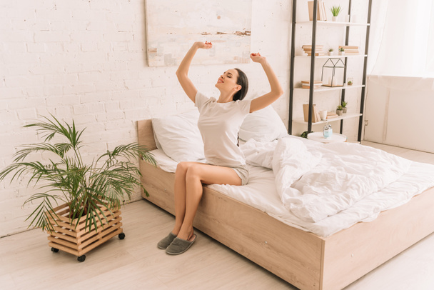 χαμογελαστή γυναίκα με πιτζάμες που τεντώνεται με κλειστά μάτια ενώ κάθεται στο κρεβάτι κοντά σε γλάστρα φυτό - Φωτογραφία, εικόνα