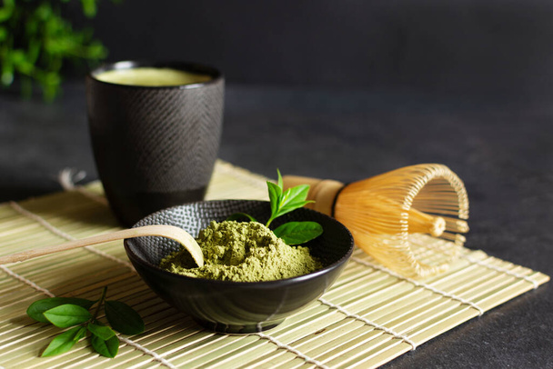 Organic green matcha tea and tea accessories on japanese mat on black background. Japanese tea ceremony concept. Chashaku spoon and chasen bamboo whisk for brewing matcha tea. - Photo, image