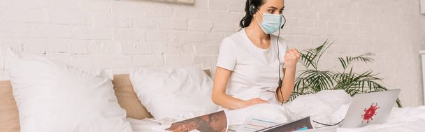 panoráma felvétel dühös távirányító headset és orvosi maszk dolgozik az ágyban, és mutatja ököl, miközben videohívás - Fotó, kép