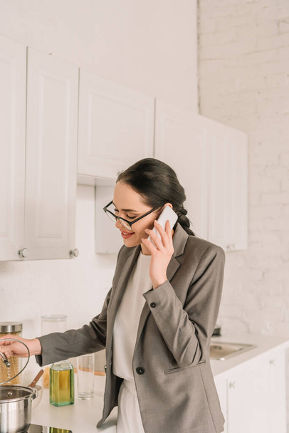 mosolygós üzletasszony blézerben pizsama felett főzés és beszélgetés okostelefonon a konyhában - Fotó, kép