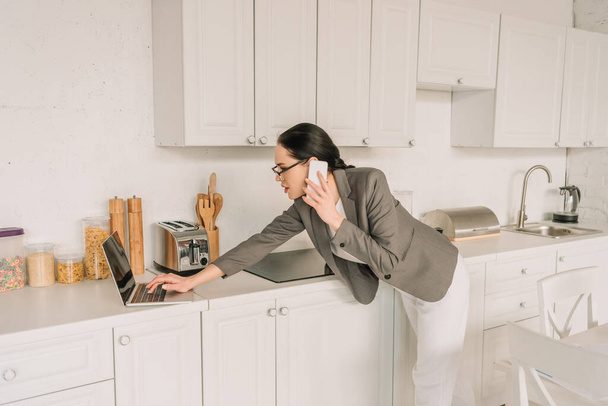 üzletasszony blézerben pizsama felett beszél okostelefonon és a laptop használata a konyhában - Fotó, kép