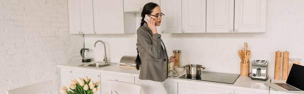 blazer ceketli bir iş kadınının mutfakta yemek pişirirken pijamalarıyla konuşmasının panoramik oryantasyonu. - Fotoğraf, Görsel