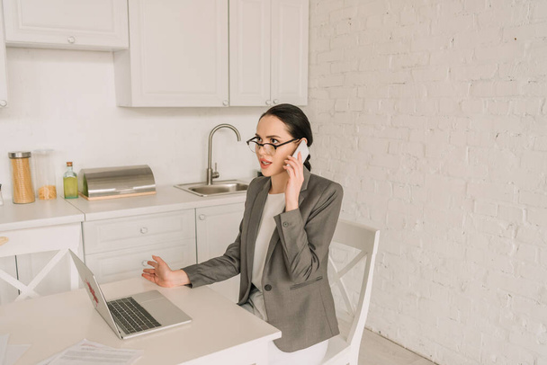 fiatal üzletasszony blézerben pizsama felett beszél okostelefonon, miközben dolgozik a konyhában laptop közelében - Fotó, kép