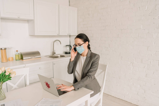 mujer de negocios en máscara médica y chaqueta sobre pijamas trabajando en la cocina, hablando en el teléfono inteligente y escribiendo en el ordenador portátil
 - Foto, imagen