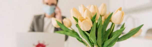 selektivní zaměření tulipánů kytice poblíž podnikatelky v lékařské masce pracuje doma, horizontální obraz - Fotografie, Obrázek