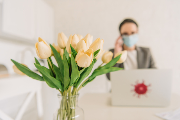 selektiver Fokus des Tulpenstraußes in der Nähe von Geschäftsfrau in medizinischer Maske, die zu Hause arbeitet - Foto, Bild