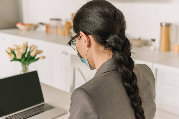 barna, hosszú hajú üzletasszony orvosi maszk dolgozik laptop a konyhában - Fotó, kép