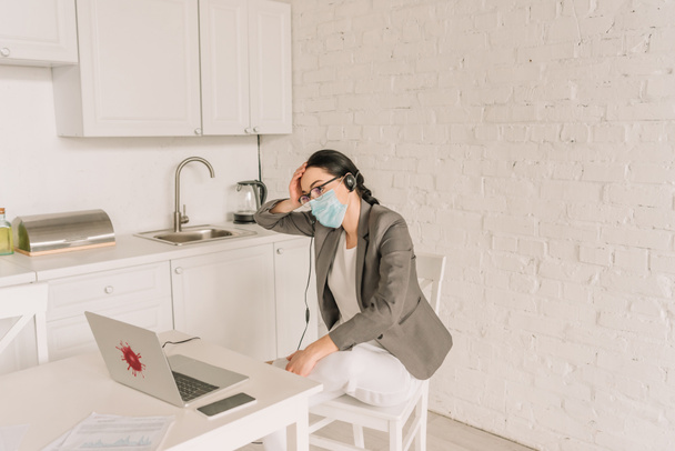 fáradt üzletasszony blézerben pizsama, orvosi maszk és headset mellett ül laptop, miközben otthon dolgozik - Fotó, kép