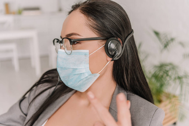 selective focus of brunette businesswoman in medical mask and eyeglasses showing victory gesture while working at home - Fotoğraf, Görsel