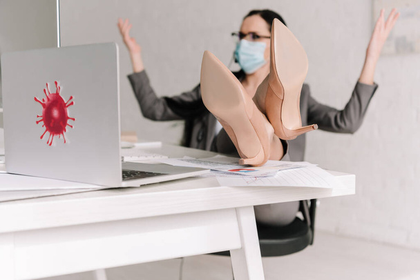 selective focus of businesswoman in medical mask showing shrug gesture while working at home with legs on table - Photo, Image