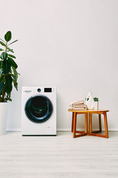 green plant near washing machine and wooden coffee table with towels and detergent bottle in bathroom  - 写真・画像