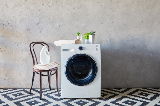 wasmiddel en spuitflessen in de buurt van wasmachine, planten, handdoeken, stoel en siertapijt in de badkamer  - Foto, afbeelding