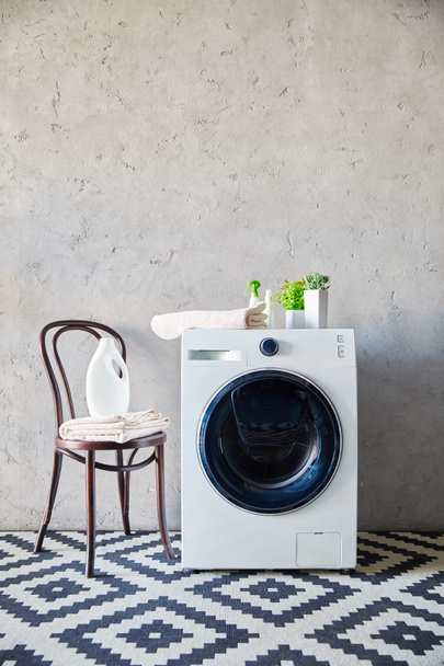 botella de detergente y toalla en la silla cerca de placas en la lavadora y alfombra ornamental en el baño
  - Foto, Imagen