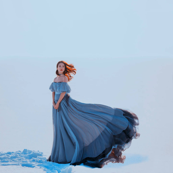 Fantasy beauty woman princess. Snow queen stands blue luxury vintage silk fabric dress fluttering in motion. Winter nature white snow. Loose red haired long hair flying in wind. lady medieval clothes - Fotó, kép