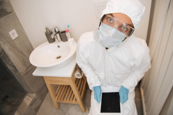 ハズマットスーツの男医療用マスクラテックスの手袋とゴーグルバスルームに空白の画面とデジタルタブレットを保持  - 写真・画像