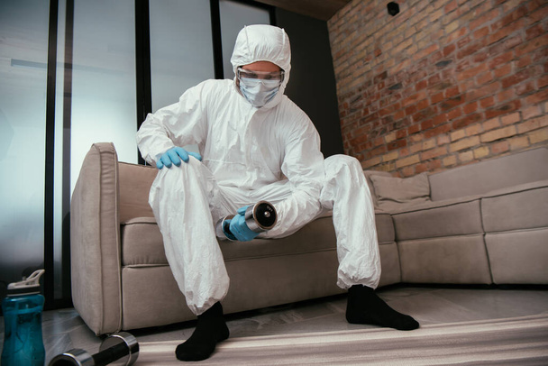 sportos férfi vegyvédelmi ruhában, orvosi maszk és védőszemüveg gyakorló súlyzóval a nappaliban  - Fotó, kép