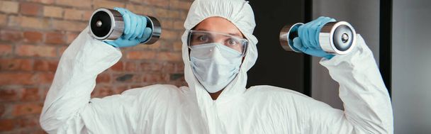 panoramic shot of sportive man in hazmat suit, medical mask and goggles exercising with dumbbells in living room  - Photo, Image