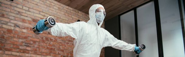 panoramic shot of sportive man in hazmat suit, medical mask and goggles working out with dumbbells in living room  - Photo, Image