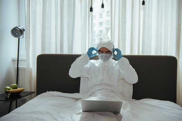 freelancer en traje de hazmat, máscara médica, guantes de látex y gafas tocando auriculares y usando laptop en el dormitorio
  - Foto, Imagen
