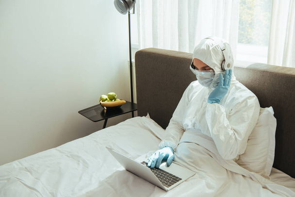 Freiberufler im Warnanzug, medizinischer Maske, Latexhandschuhen und Brille, die Kopfhörer berühren und Laptop im Bett benutzen - Foto, Bild