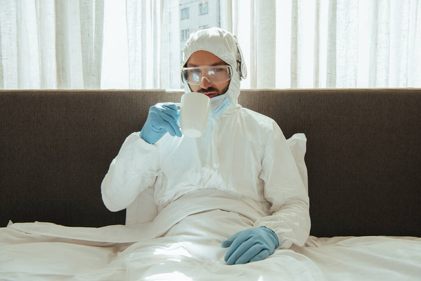 Mann im Warnanzug, Kopfhörer, medizinische Maske, Latexhandschuhe und Schutzbrille im Schlafzimmer  - Foto, Bild