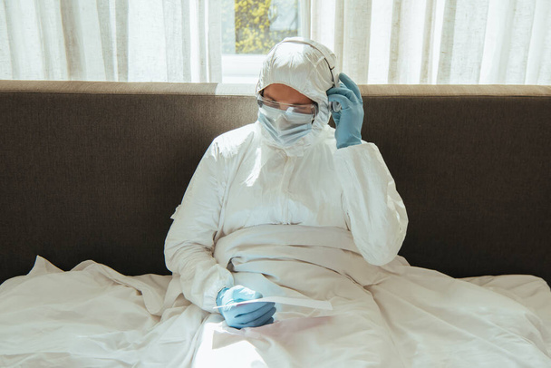 man in personal protective equipment holding paper and touching headphones in bedroom  - Photo, Image