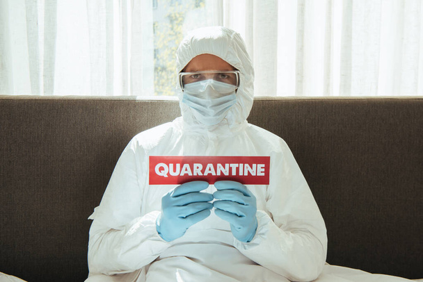 Mann in persönlicher Schutzausrüstung hält Papier mit Quarantäne-Schriftzug im Schlafzimmer  - Foto, Bild