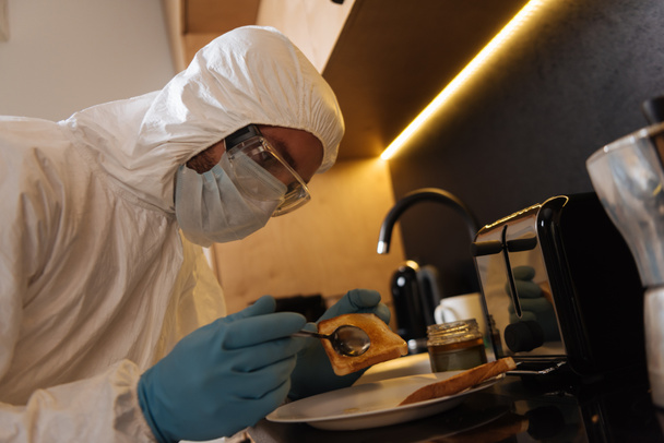 άνθρωπος σε μέσα ατομικής προστασίας κρατώντας κουτάλι κοντά τοστ ψωμί στην κουζίνα  - Φωτογραφία, εικόνα