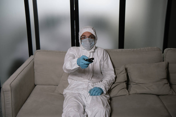 άνθρωπος σε ατομικό προστατευτικό εξοπλισμό κρατώντας τηλεχειριστήριο, ενώ βλέποντας ταινία στο σαλόνι  - Φωτογραφία, εικόνα