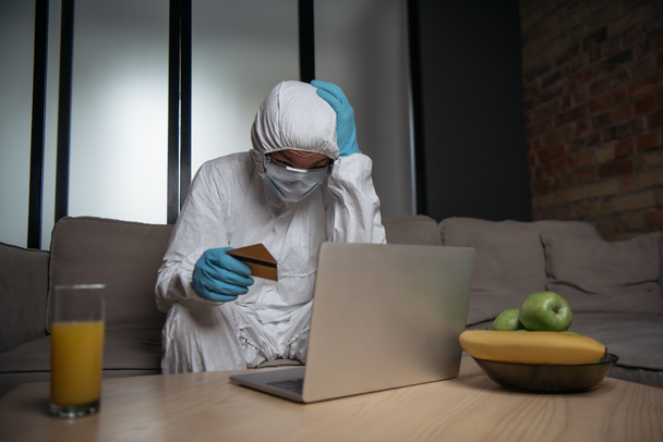 mężczyzna w osobistym sprzęcie ochronnym i masce medycznej za pomocą laptopa i trzymając karty kredytowe w pobliżu owoców i szklanki soku pomarańczowego  - Zdjęcie, obraz