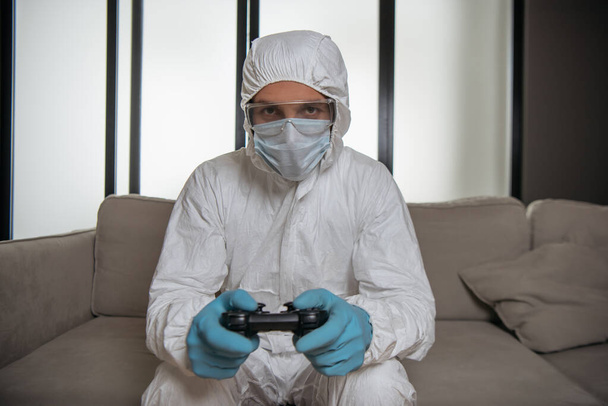 KYIV, UKRAINE - APRIL 11, 2020: man in personal protective equipment and medical mask playing video game  - Photo, Image
