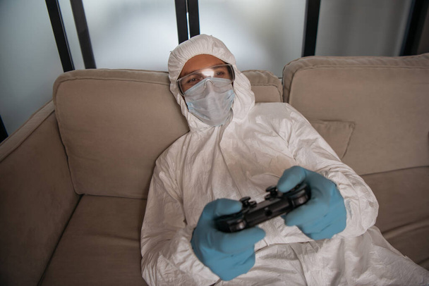 KYIV, UKRAINE - APRIL 11, 2020: man in personal protective equipment and medical mask playing video game in living room  - Photo, image