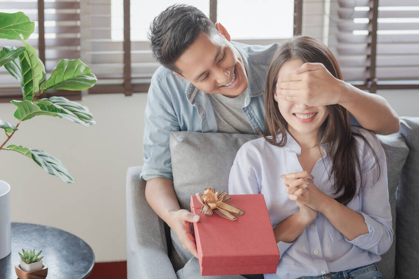 Όμορφος άντρας δίνει στην κοπέλα του ένα κόκκινο κουτί δώρου για γενέθλια έκπληξη στο σαλόνι του σπιτιού. - Φωτογραφία, εικόνα