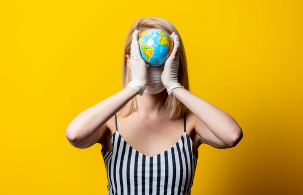 Blonde woman in face mask and gloves holds Earth globe on yellow background - Photo, Image