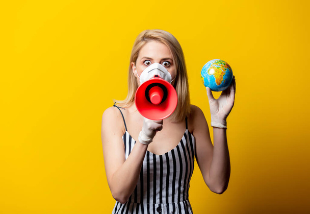 Blonde woman in face mask and gloves holds Earth globe and megaphone on yellow background - Foto, Imagen