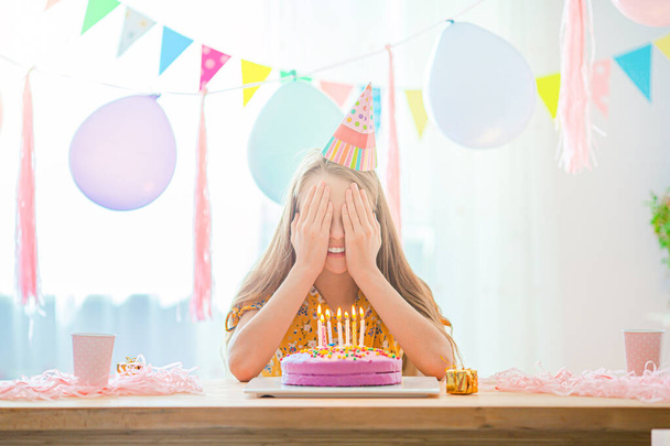 Caucasian girl is dreamily smiling and looking at birthday rainbow cake. Festive colorful background with balloons. Birthday party and wishes concept. - Photo, Image