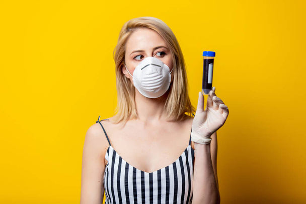 Blonde woman in face mask and gloves holds jar with blood on yellow background - Photo, Image