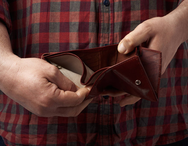 man in a red plaid shirt holds an empty leather brown wallet, concept of poverty and economic crisis - Photo, Image
