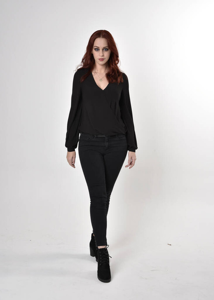 Portrait of a pretty girl with red hair wearing black jeans, boots and a blouse.  full length standing pose, facing the camera, on a studio background. - Fotografie, Obrázek