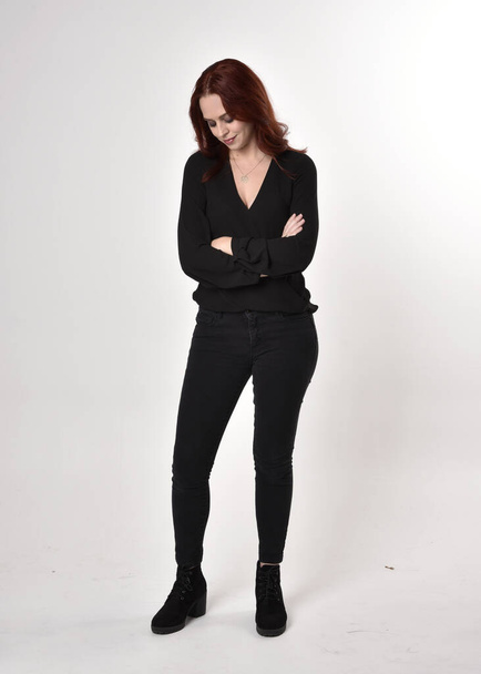 Портрет красивой девушки с рыжими волосами в черных джинсах, сапогах и блузке. полная поза стоя, лицом к камере, на фоне студии
. - Фото, изображение