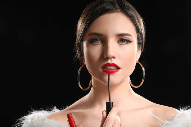 Beautiful young woman applying lipstick against dark background - Foto, afbeelding