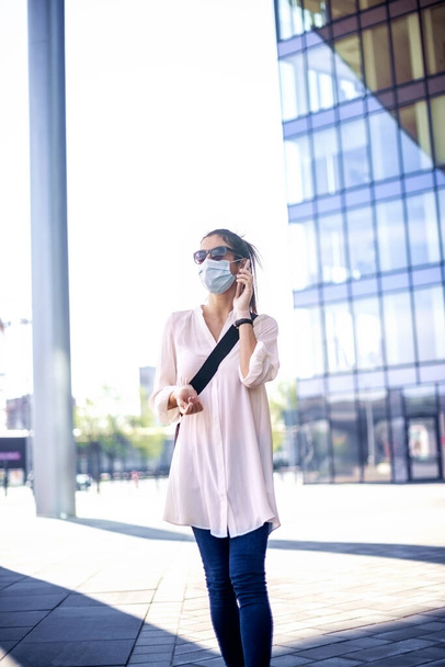 Full length shot of confident woman wearing face mask and talking with somebody on her mobile phone while walking in front of business center during coronavirus pandemic. - 写真・画像